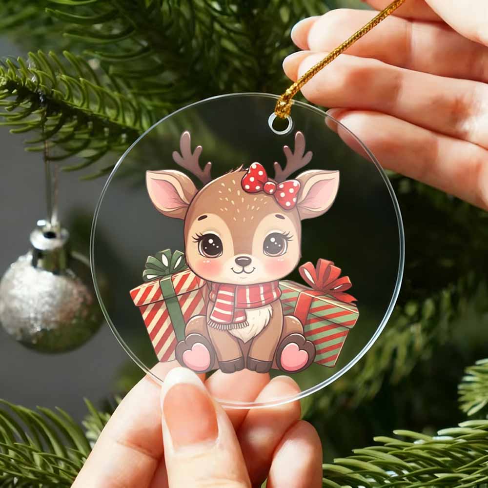 Clear Christmas ornament with a cartoon deer holding presents, held by hands against a Christmas tree background.