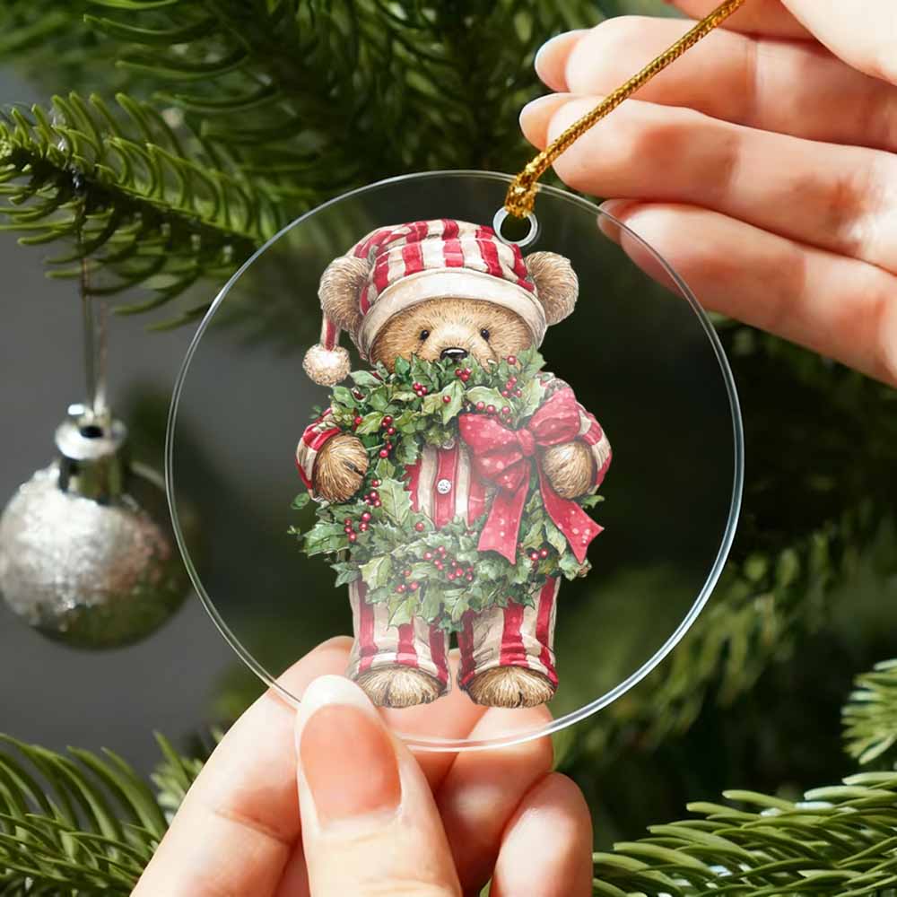 Christmas ornament with a teddy bear holding a wreath, held by hands against a Christmas tree background.
