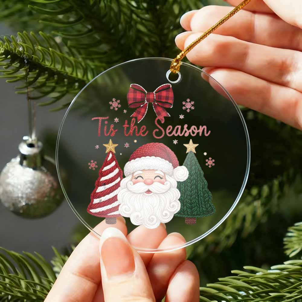 Christmas ornament with Santa Claus and text 'Tis the Season' held by a hand against a Christmas tree background.